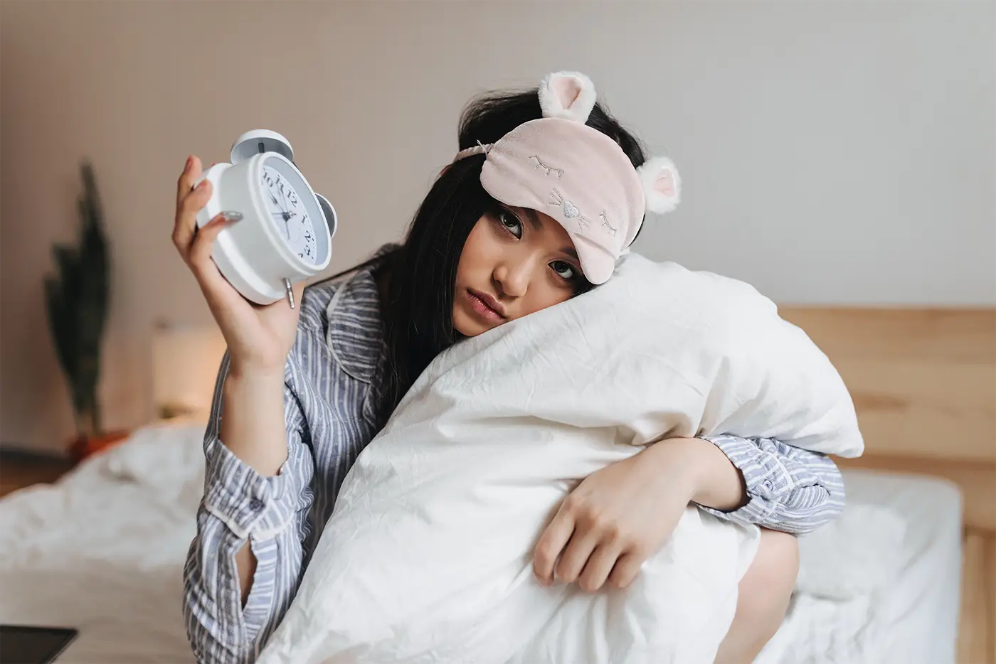 sad-girl-pajamas-sleep-mask-is-hugging-white-pillow-holding-alarm-clock
